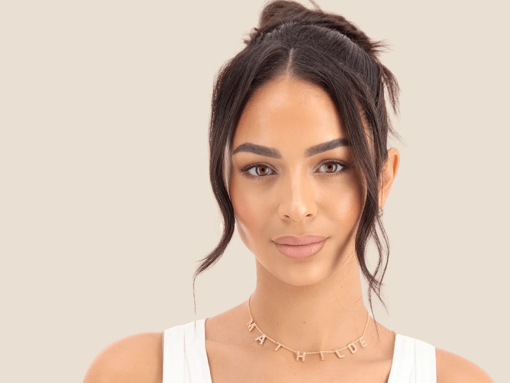 Femme aux cheveux bruns relevés, top blanc et collier doré orné de lettres, sur fond beige